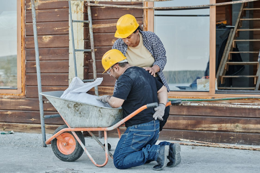 Two construction workers addressing a task with a wheelbarrow, emphasising gaps in workplace injury management, such as lack of proper ergonomics and safety oversight on construction sites.